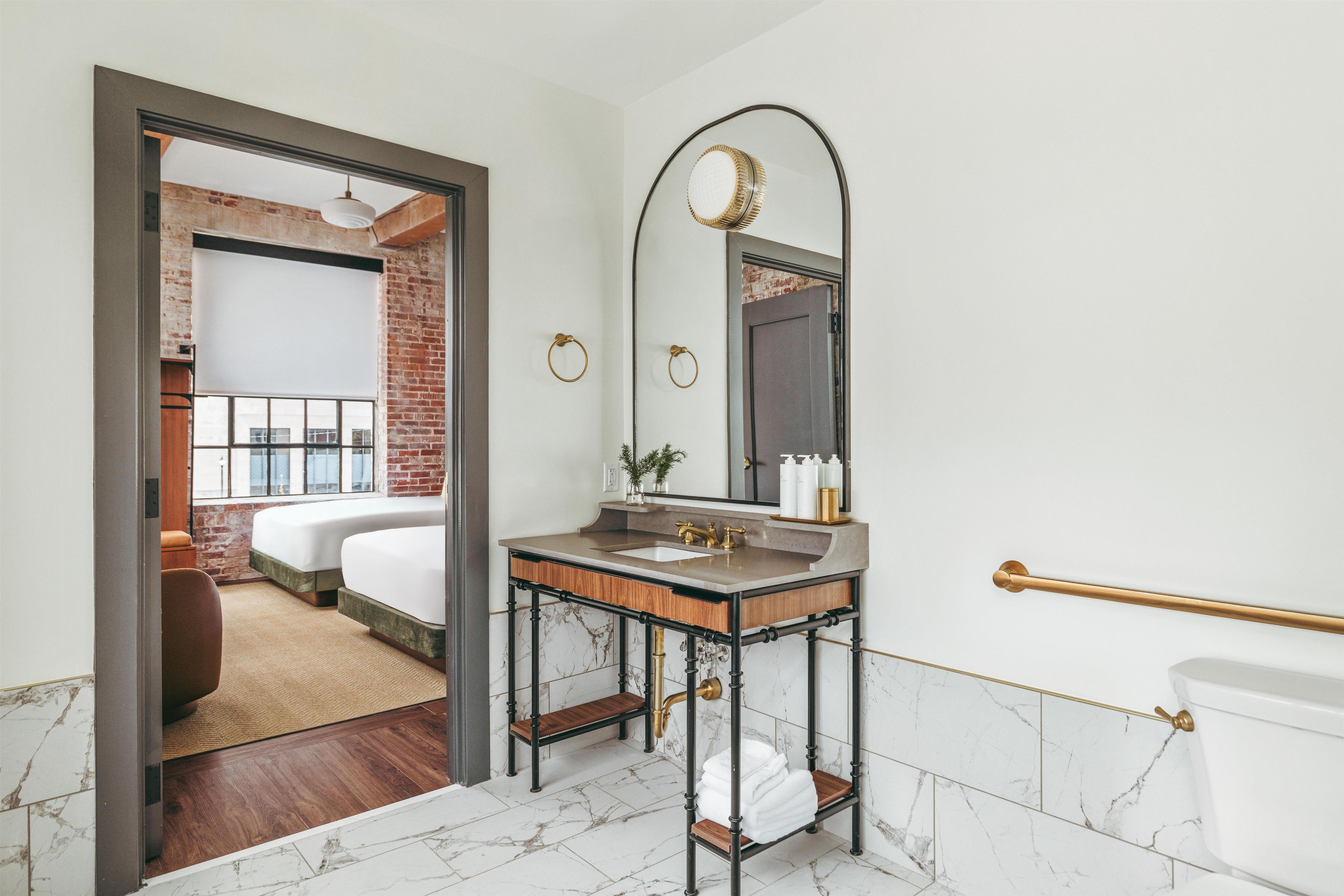 A modern bathroom with a marble floor, a wooden vanity, a large mirror, and a toilet with a gold handle.
