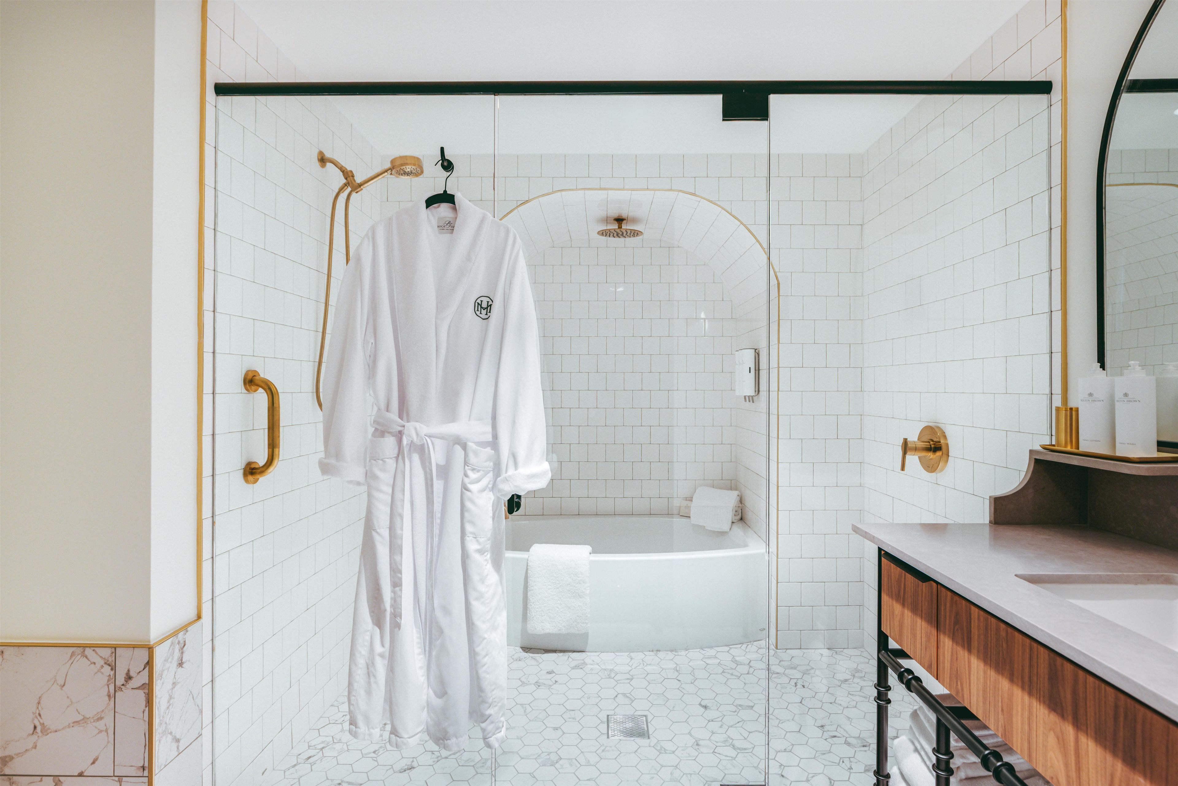 Une salle de bain moderne avec des carreaux de céramique blancs rectangulaires, une baignoire blanche, un meuble-lavabo en bois et un peignoir suspendu à un porte-serviettes.