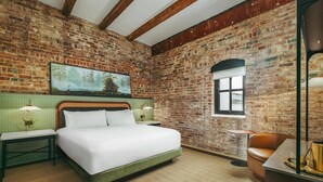 A room with a brick wall, a bed with white bedding, a green headboard, a painting, a window, and a brown leather chair. - The Hotel Monroe, Tapestry Collection By Hilton (Monroe)