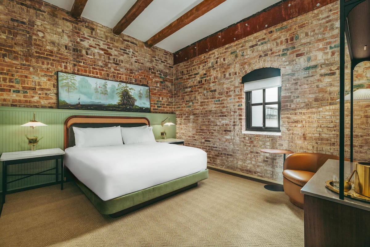 Una habitación con pared de ladrillo, una cama con ropa blanca, cabecera verde, un cuadro, una ventana y un sillón de cuero marrón.