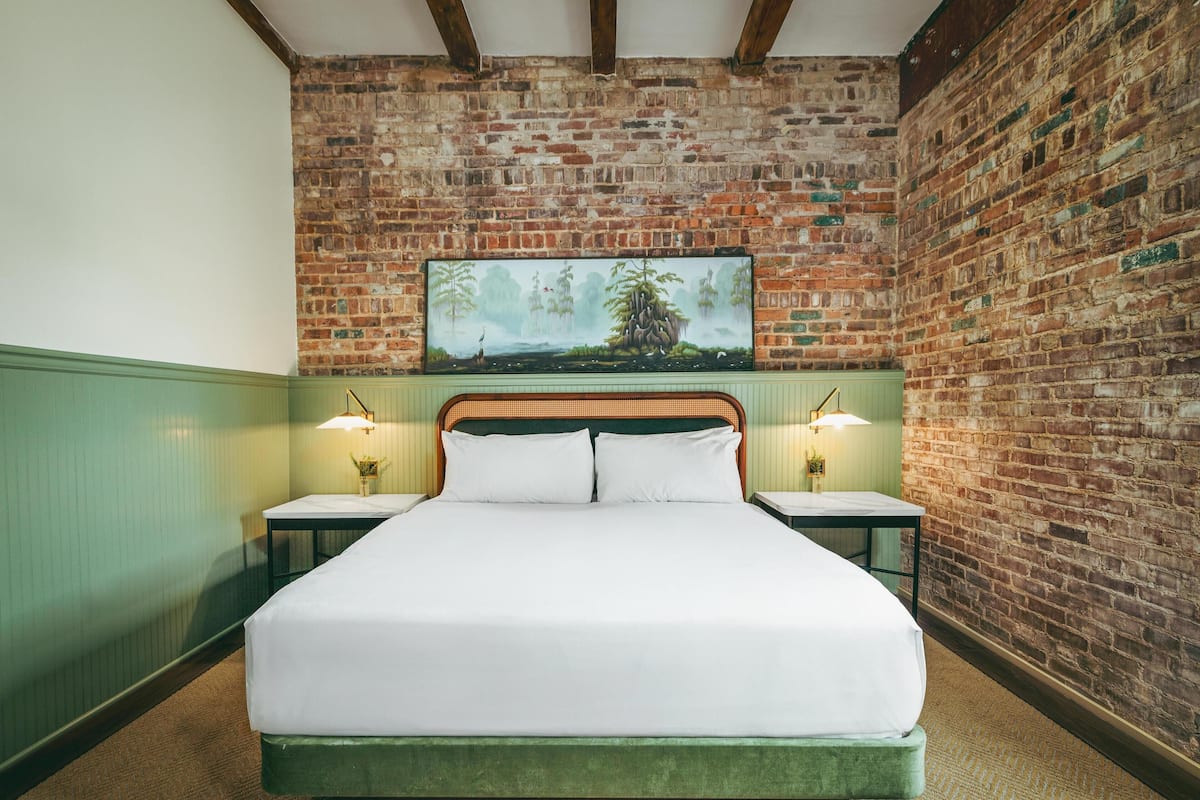 Una cama bien hecha en una habitación con pared de ladrillo y un cuadro colgado. Hay dos mesitas de noche con lámparas.