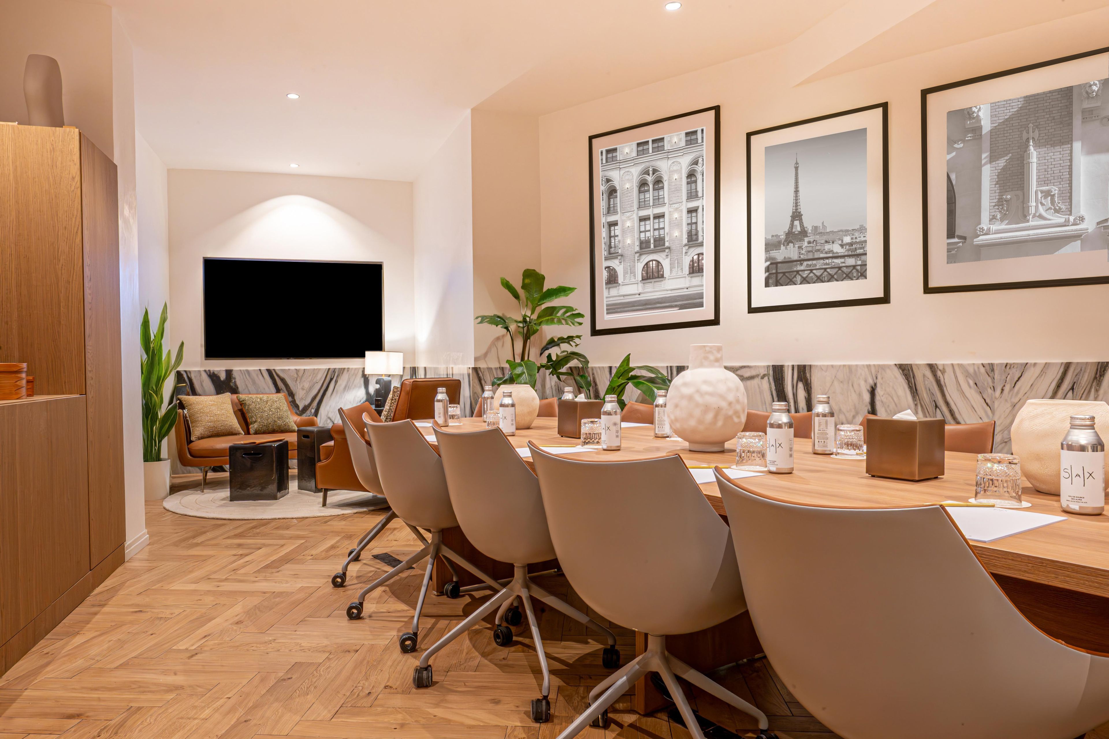 A modern hotel room with a large flat-screen TV, a dining area with a wooden table and chairs, and framed pictures on the wall.