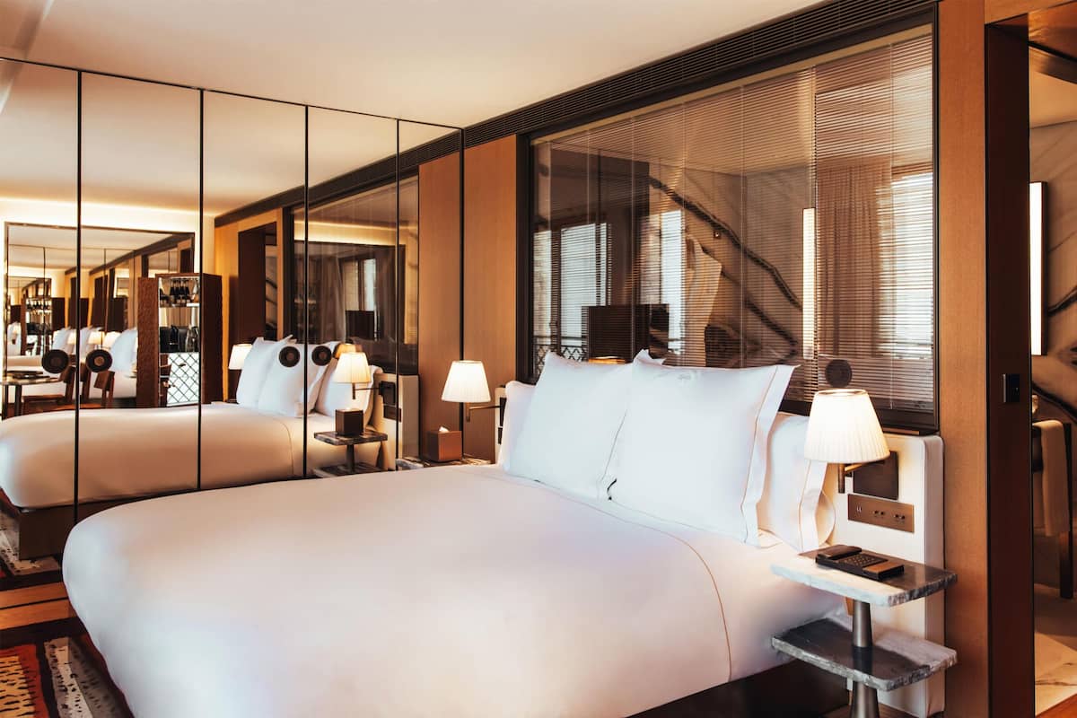 Moderni hotellihuone, jossa on suuri sänky, yöpöydät, lamput ja suuri ikkuna, jossa on sälekaihtimet.