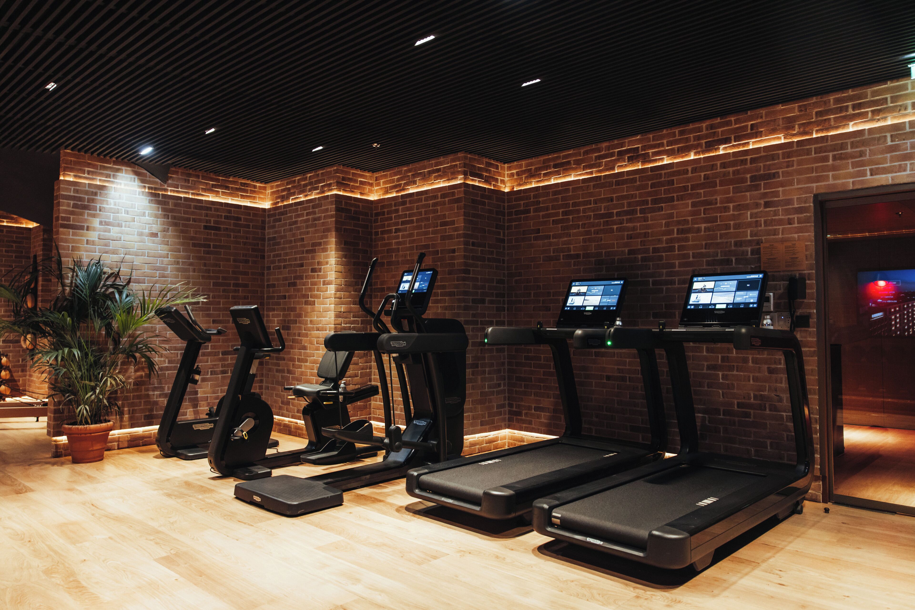 A gym with treadmills, elliptical machines, and a stationary bike, featuring brick walls and wooden flooring.