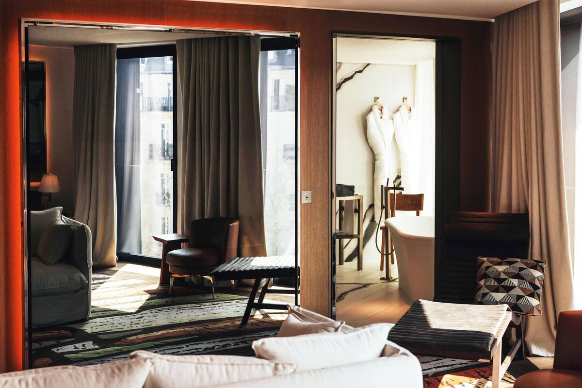 Hotellihuone, jossa on sänky, sohva, tuoli, pieni pöytä ja näkymä avoimesta ovesta.