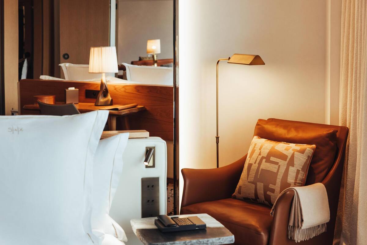 Hotellihuone, jossa on sänky, kirjoituspöytä lampun kanssa, nahkainen nojatuoli ja yöpöytä, jossa on kaukosäädin.