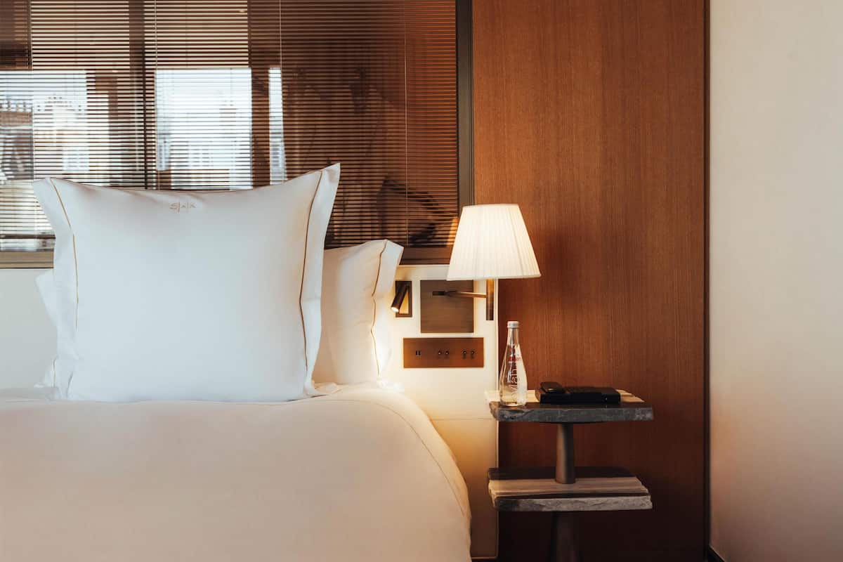Hotellihuone, jossa on sänky, yöpöytä lampun kanssa, pullo ja puhelin.