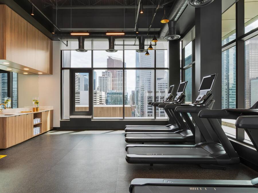 A modern gym with treadmills, a wooden counter, and large windows offering city views.