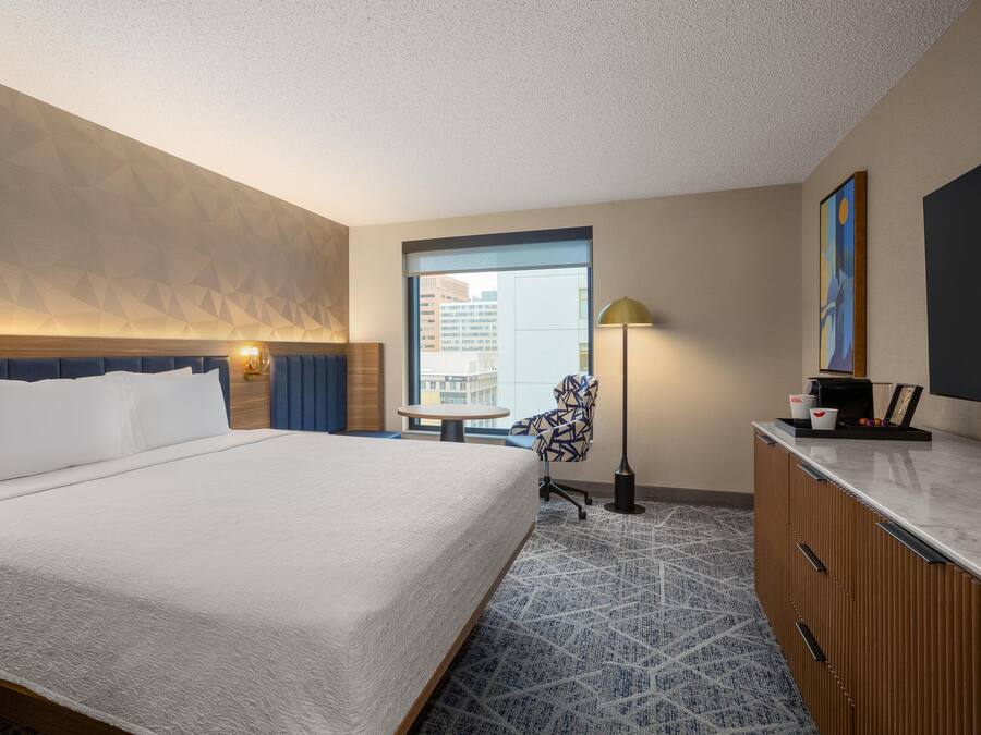 Habitación de hotel con una cama grande, un escritorio, una silla, una lámpara y vistas a la ciudad a través de una ventana.