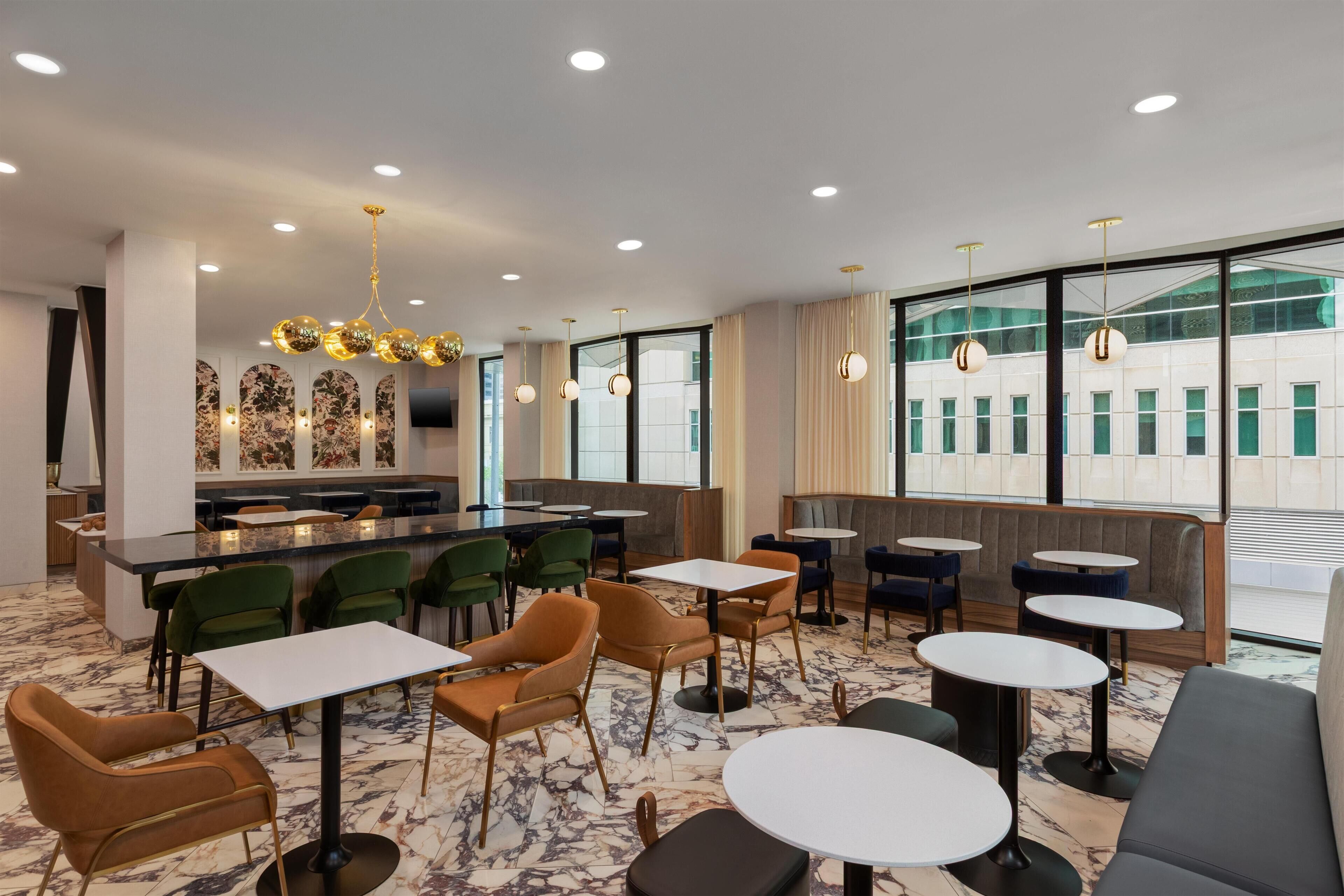 A modern restaurant with marble flooring, green and brown chairs, round tables, and a bar area with artwork on the wall.