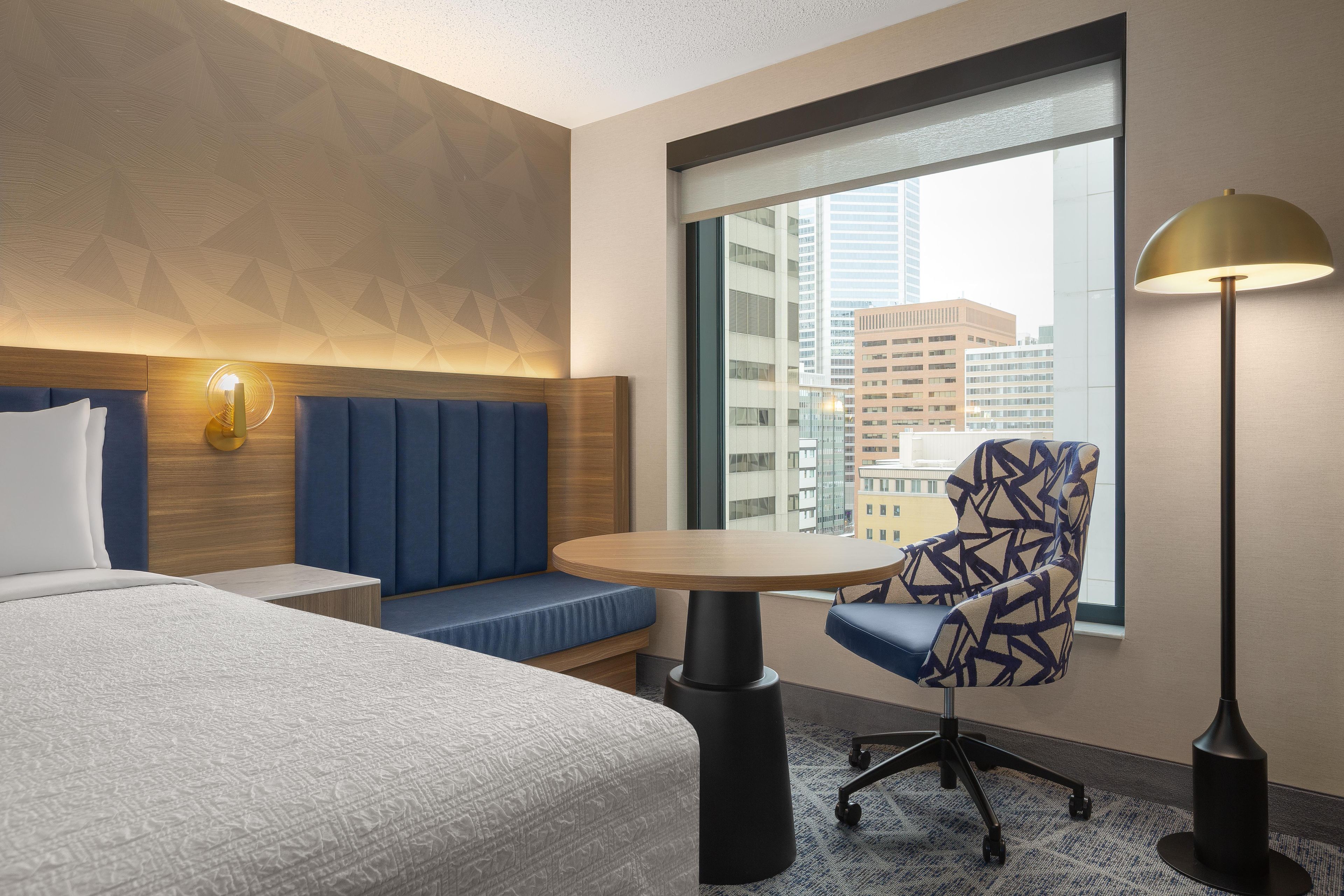 Quarto de hotel com cama, área de estar com mesa, luminária e vista para a cidade através de uma janela grande.