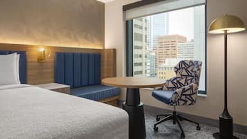 Quarto de hotel com cama, área de estar com mesa, luminária e vista para a cidade através de uma janela grande.
