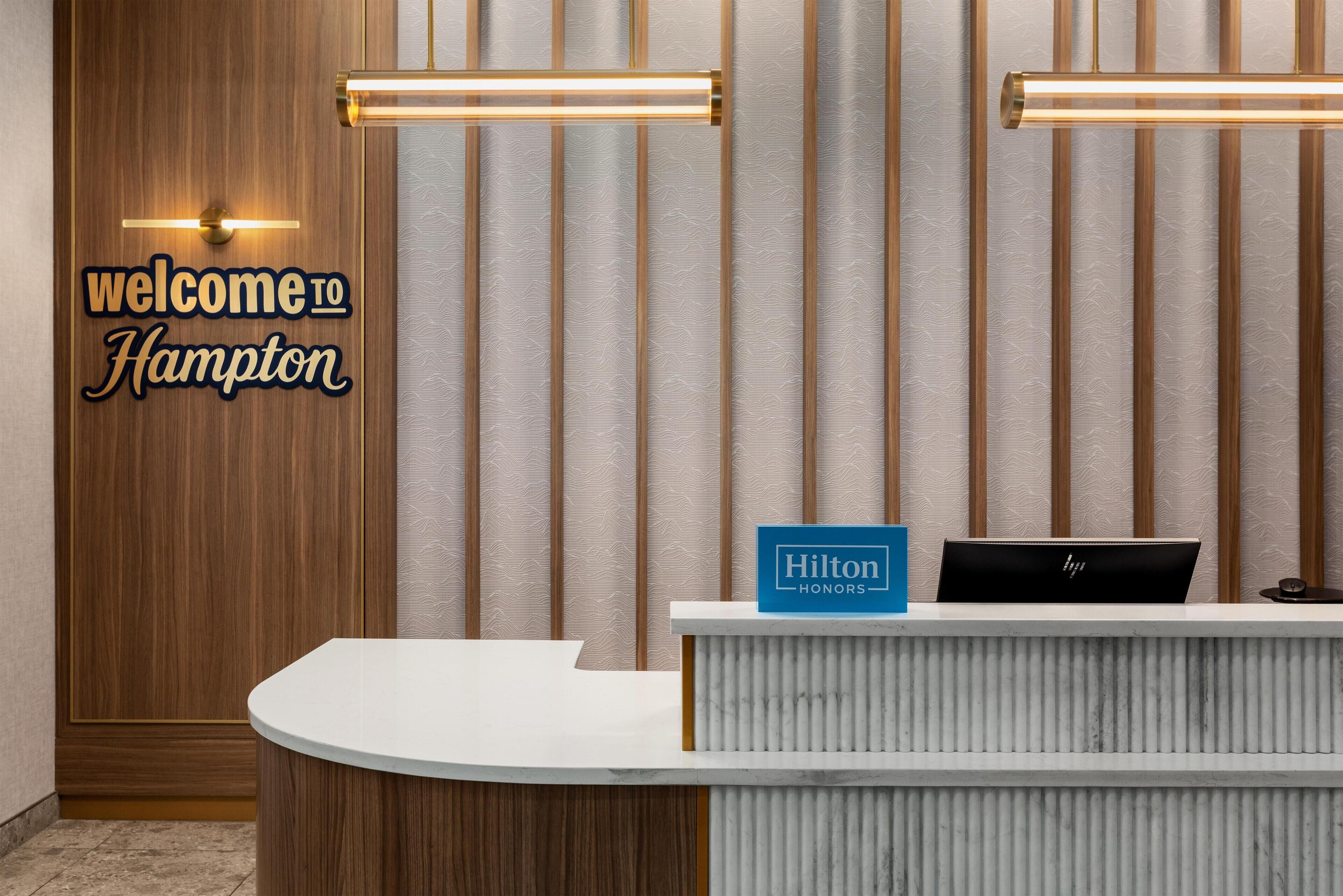 Un mostrador de recepción de hotel con un cartel que dice 'welcome to Hampton', un cartel de 'Hilton Honors' y una computadora portátil negra sobre el mostrador.