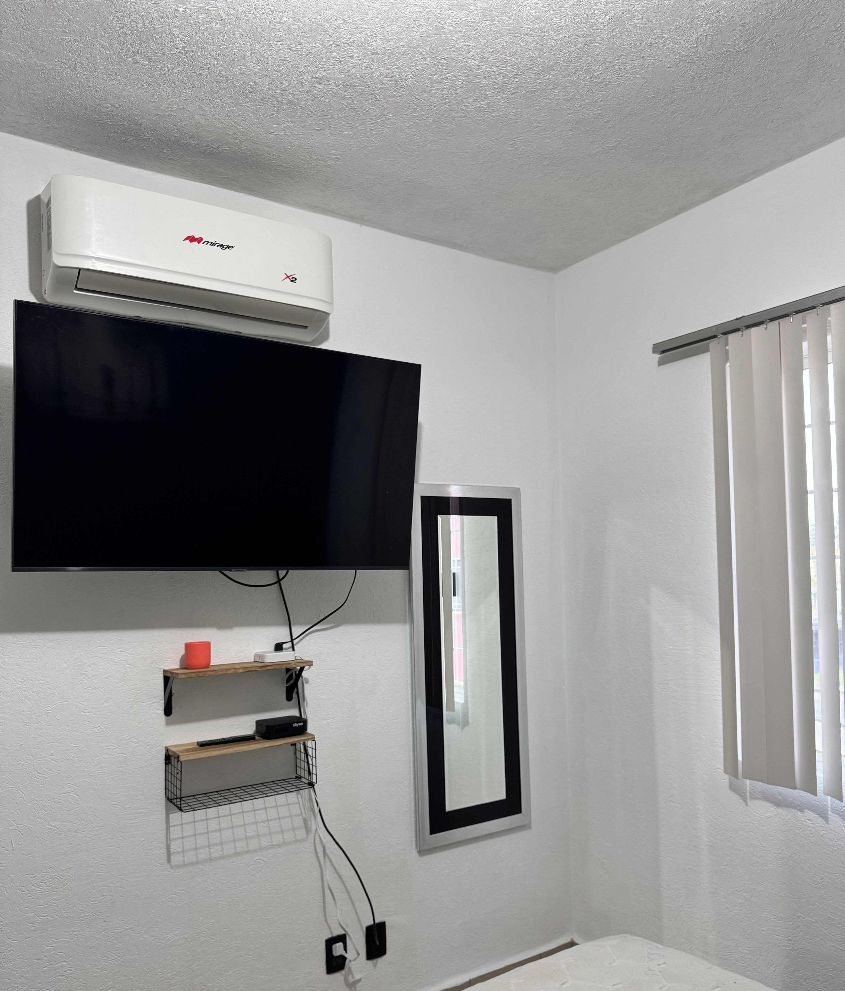 Een kamer met een ingebouwde airconditioner, een flatscreen televisie, een spiegel en een raam met jaloezieën.