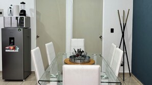 A modern dining area with a glass table, white chairs, a clock, and a refrigerator with a coffee maker.