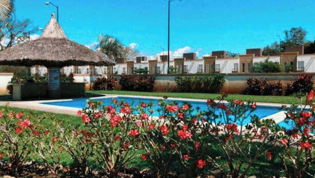 A pool surrounded by red flowers, a thatched-roof structure, and a row of white buildings.