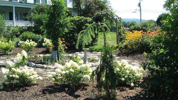 Un jardin agrémenté de diverses plantes et fleurs, un sentier en pierre et, au fond, une maison blanche.