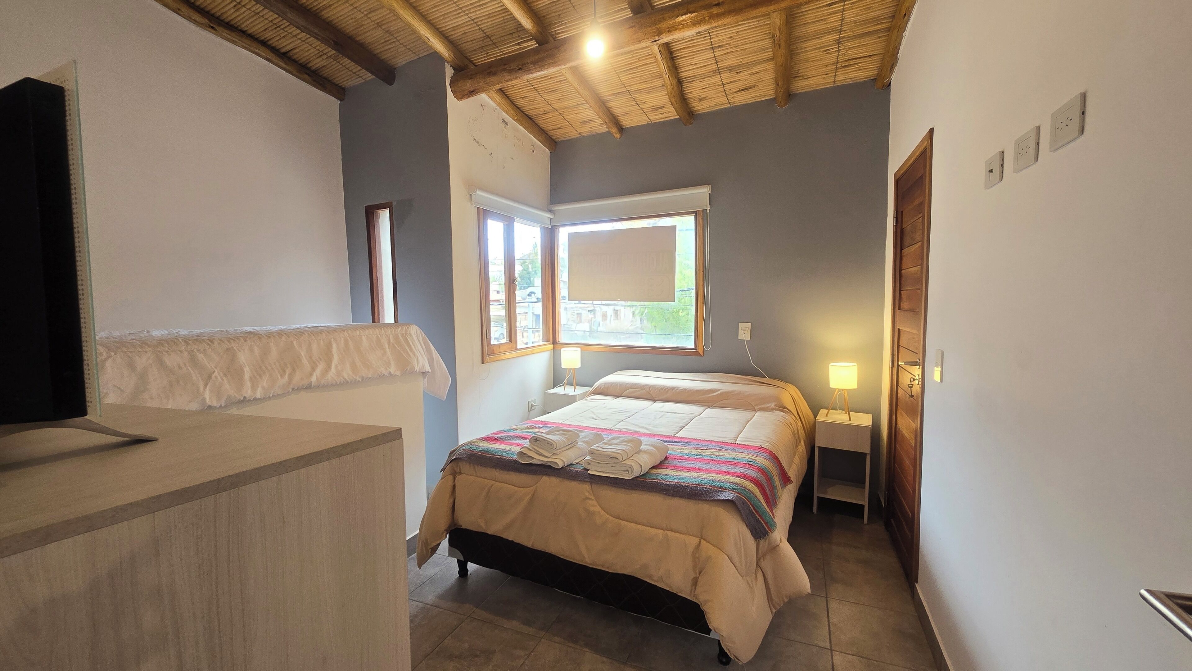 A bedroom with a bed, bedside tables, a window, and a wooden ceiling.