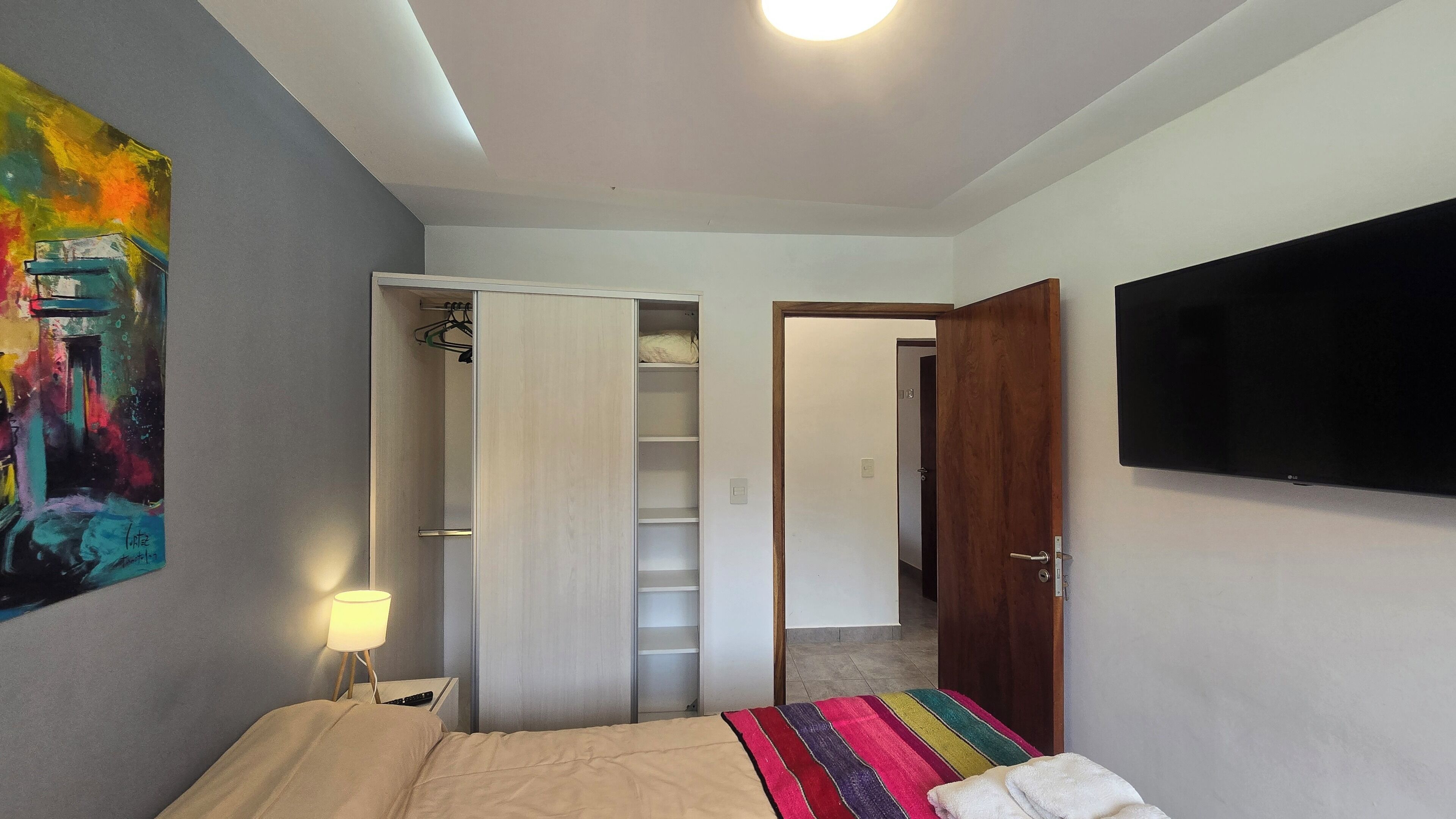 A bedroom with a bed, a TV mounted on the wall, a wardrobe, and a colorful painting on the wall.