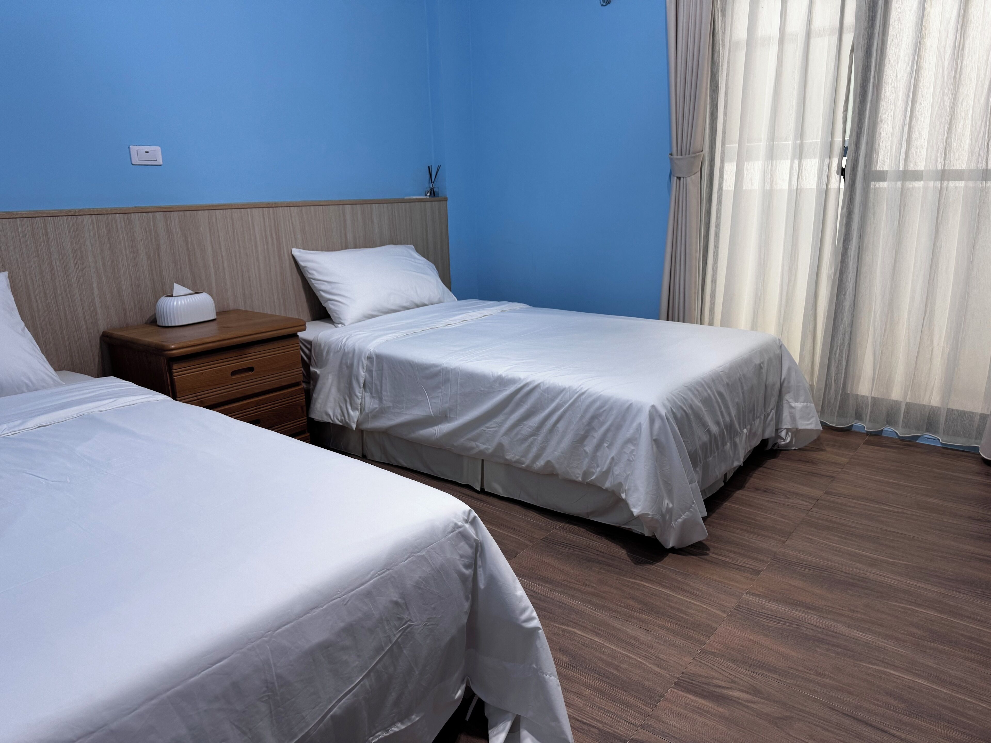 A hotel room with two beds, wooden headboard, a nightstand, and a window with sheer curtains.