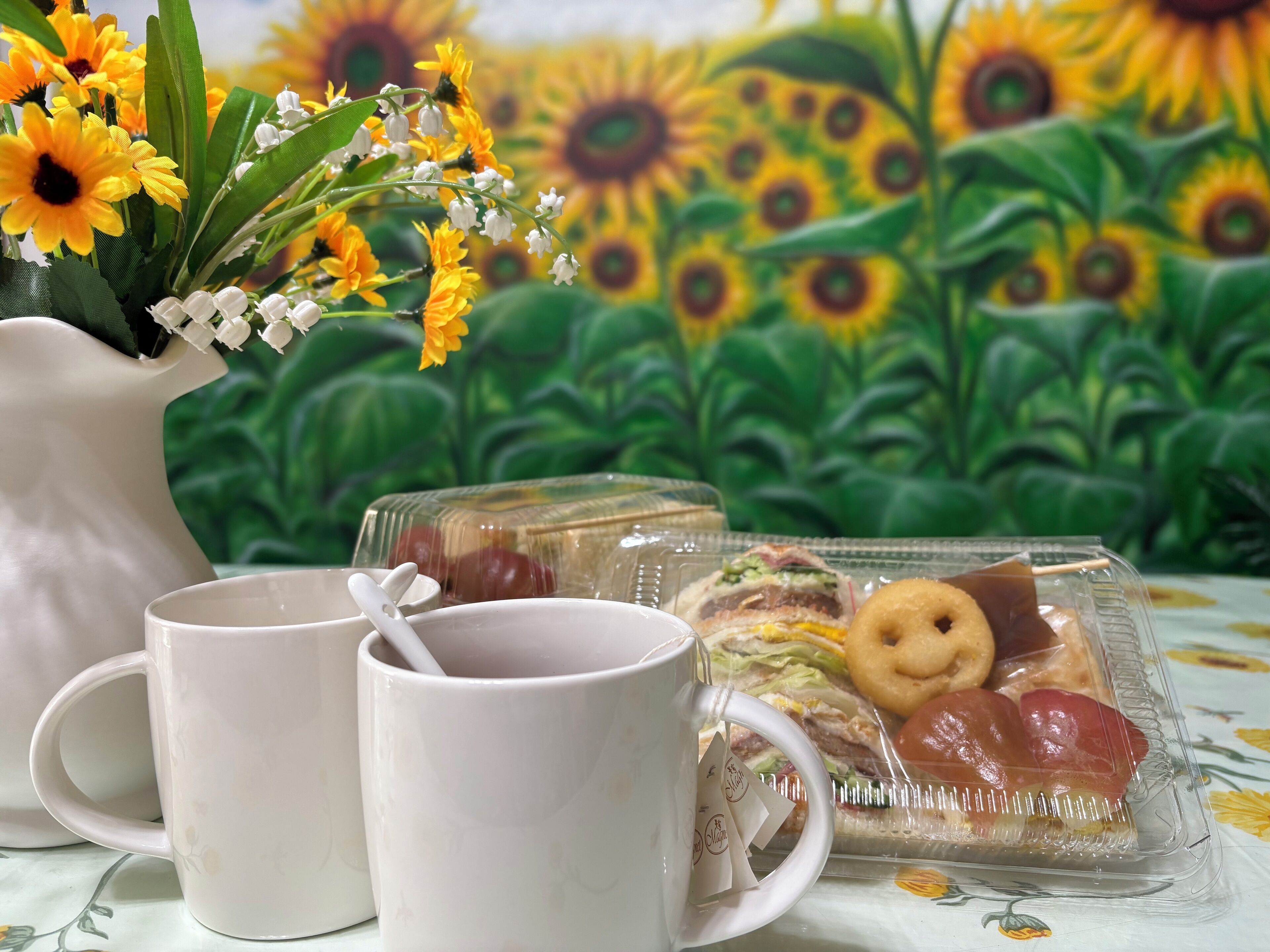 A table with two white mugs, a clear plastic tray with sandwiches and a biscuit, and a white pitcher with flowers.
