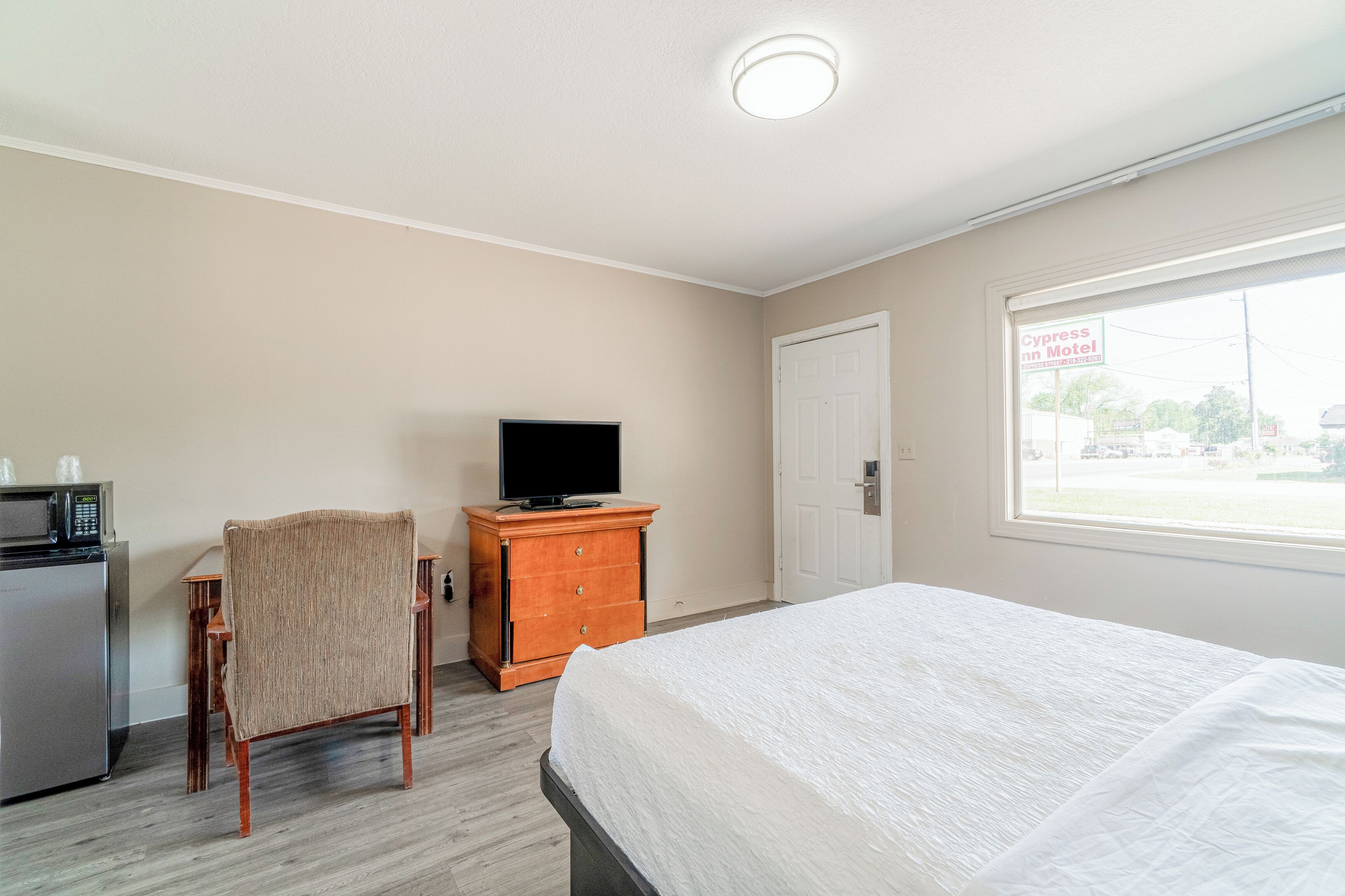 A hotel room with a bed, a TV on a wooden dresser, a microwave, a chair, and a window with a view of the outside.