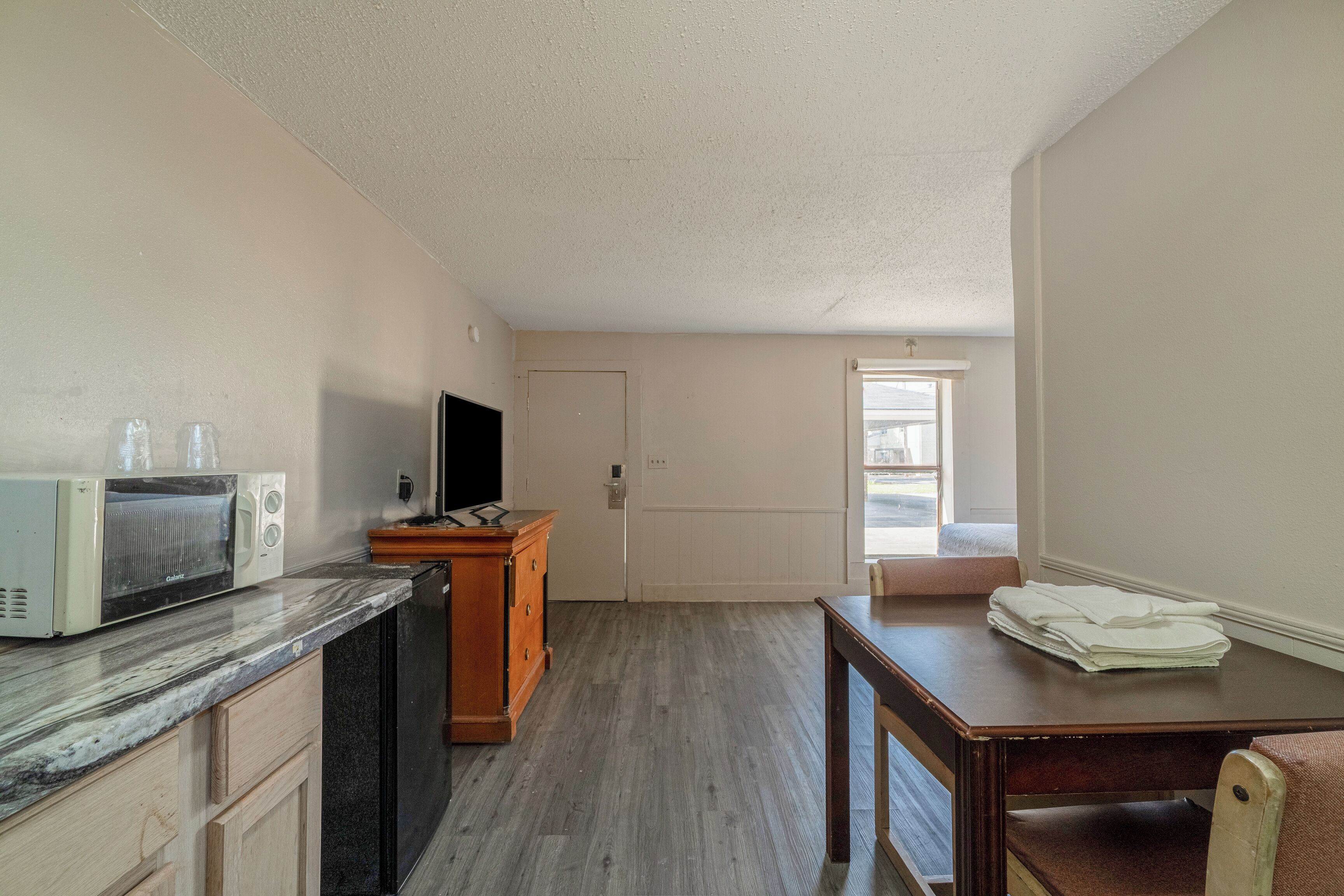 A kitchen with a microwave, wooden cabinet, and a television. There is a door leading to another room and a window with natural light.