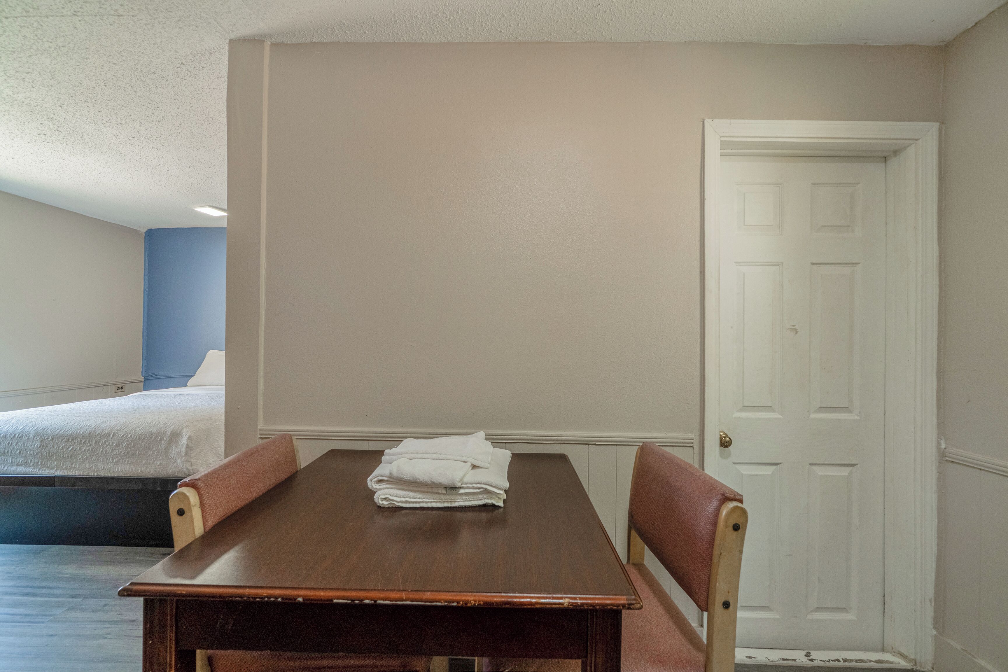 Una habitación con una mesa de madera, dos sillas, una cama y una puerta blanca.