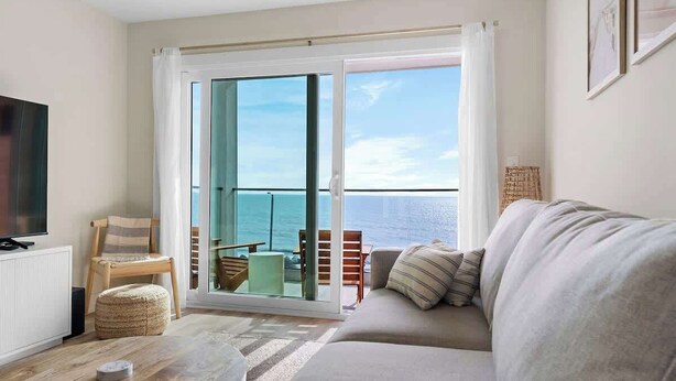 A living room with a sofa, a television, a coffee table, and a view of the sea through a sliding glass door. - Oceanside Oasis by Carolina Getaway - Oceanfront Condo POOL & HOT TUB OPEN (Carolina Beach)