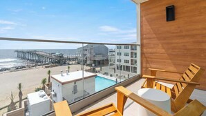 A balcony with a view of the beach and pier, wooden chairs, a small pool, and a modern building in the background. - Oceanside Oasis by Carolina Getaway - Oceanfront Condo POOL & HOT TUB OPEN (Carolina Beach)