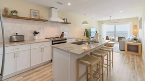 A modern kitchen with white cabinetry, a central island with stools, a large window, and a view of the sea. - Oceanside Oasis by Carolina Getaway - Oceanfront Condo POOL & HOT TUB OPEN (Carolina Beach)