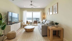 A modern living room with a flat-screen TV, a ceiling fan, a sofa, a coffee table, and a balcony with a view. - Oceanside Oasis by Carolina Getaway - Oceanfront Condo POOL & HOT TUB OPEN (Carolina Beach)