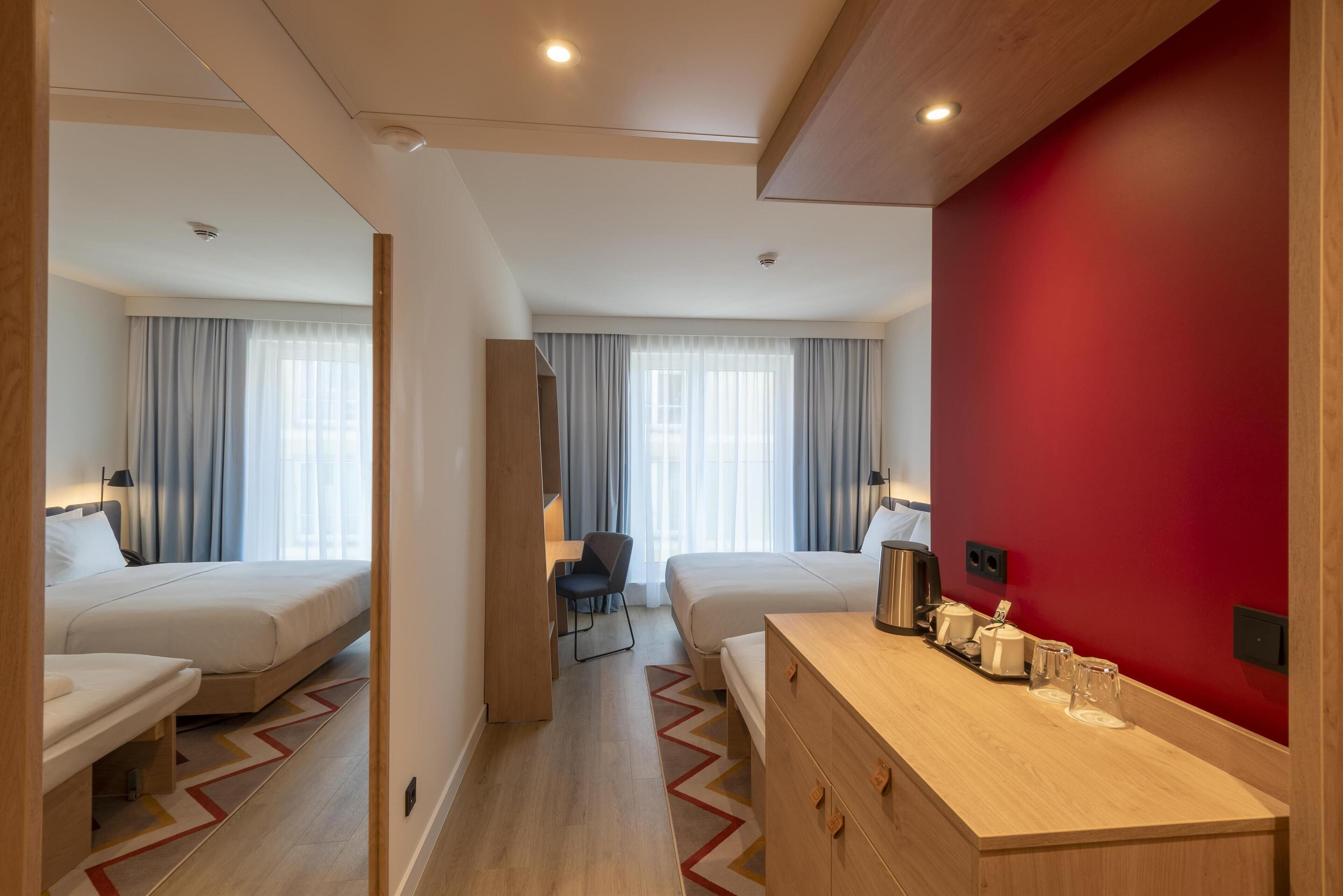 A hotel room with two beds, a desk, a chair, a red accent wall, and a wooden dresser with a tray of glasses.