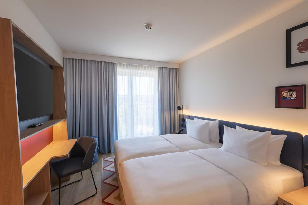 Une chambre d’hôtel avec deux lits, un bureau, une chaise, une télévision et une fenêtre avec des rideaux.