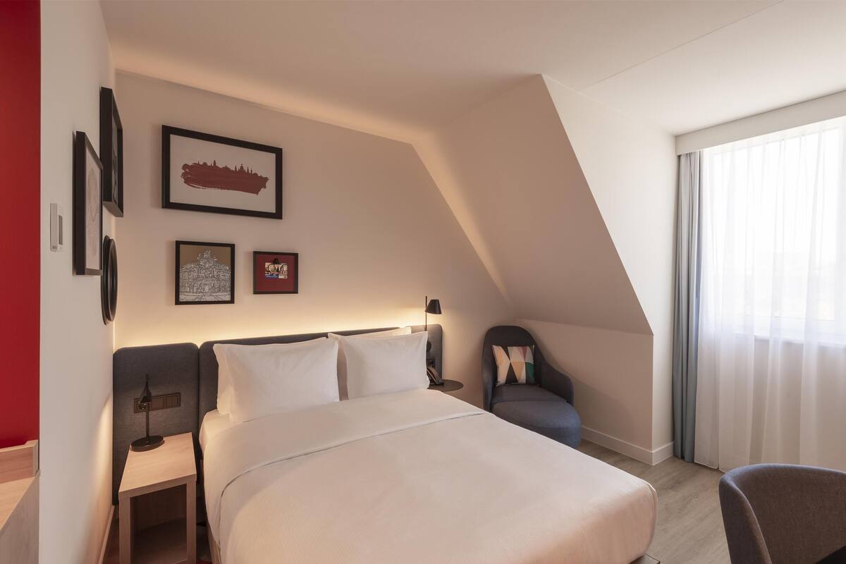 Une chambre d’hôtel moderne avec un grand lit, un bureau et une chaise. Les murs sont ornés de tableaux encadrés et il y a une fenêtre avec des rideaux.