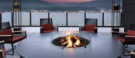 A round table with a fire pit in the center, surrounded by chairs, overlooking a body of water and distant mountains.