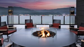 A round table with a fire pit in the center, surrounded by chairs, with a view of a body of water and mountains in the background. - Tempo By Hilton Pigeon Forge (Pigeon Forge)