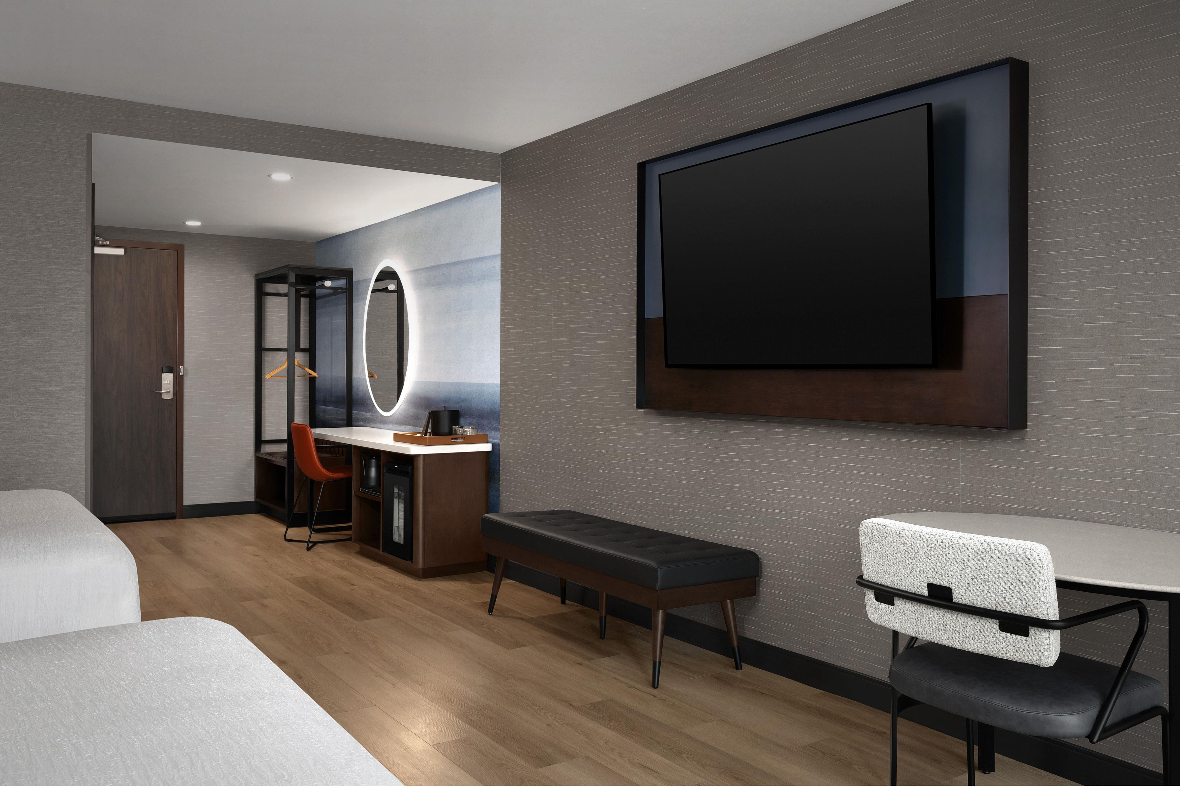 Ein modernes Hotelzimmer mit einem großen Bett, einem Schreibtisch mit Stuhl, einer Bank, einem an der Wand montierten Fernseher und einem Spiegel.