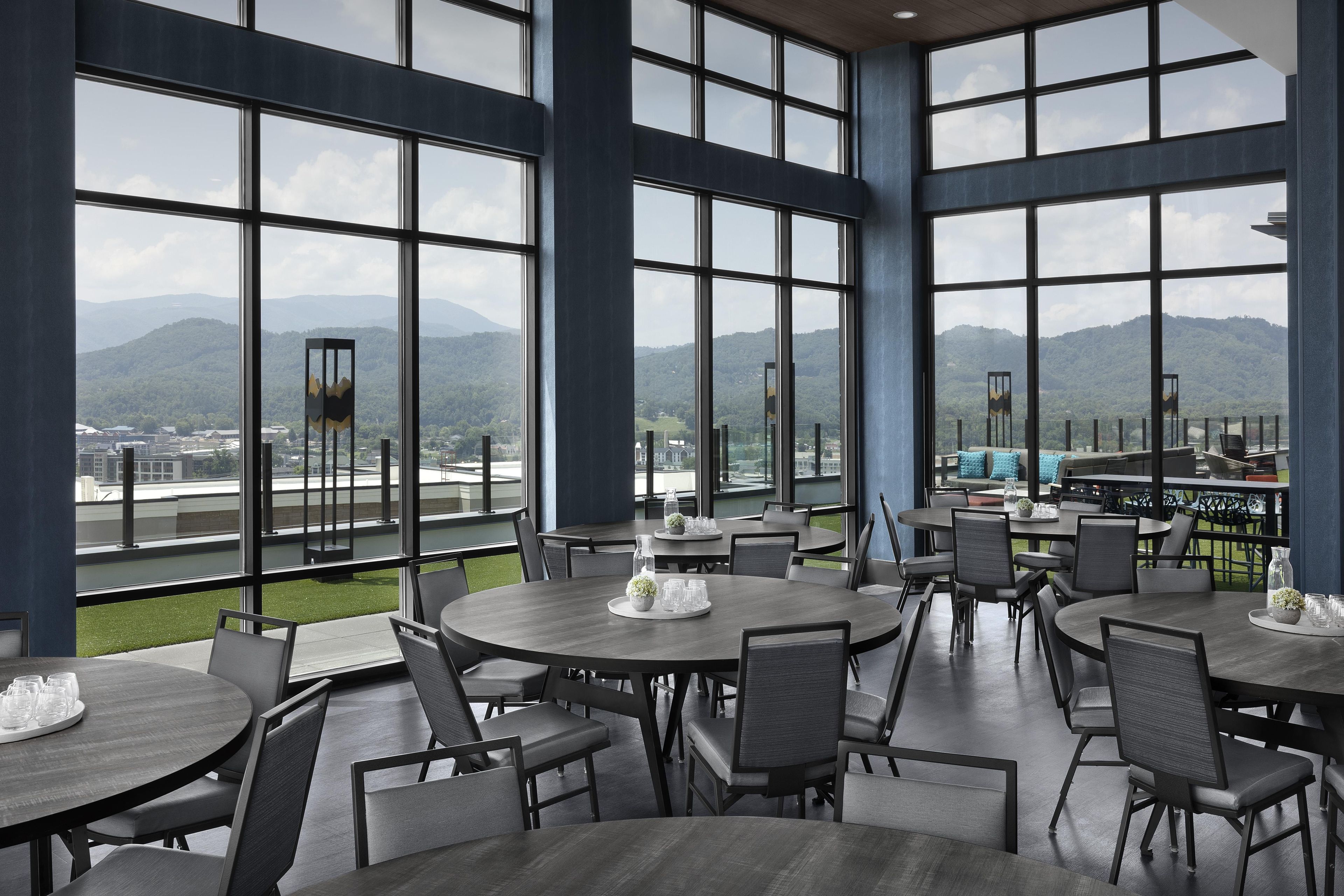 A restaurant with large windows offering a view of a city and mountains. Tables are arranged for dining.