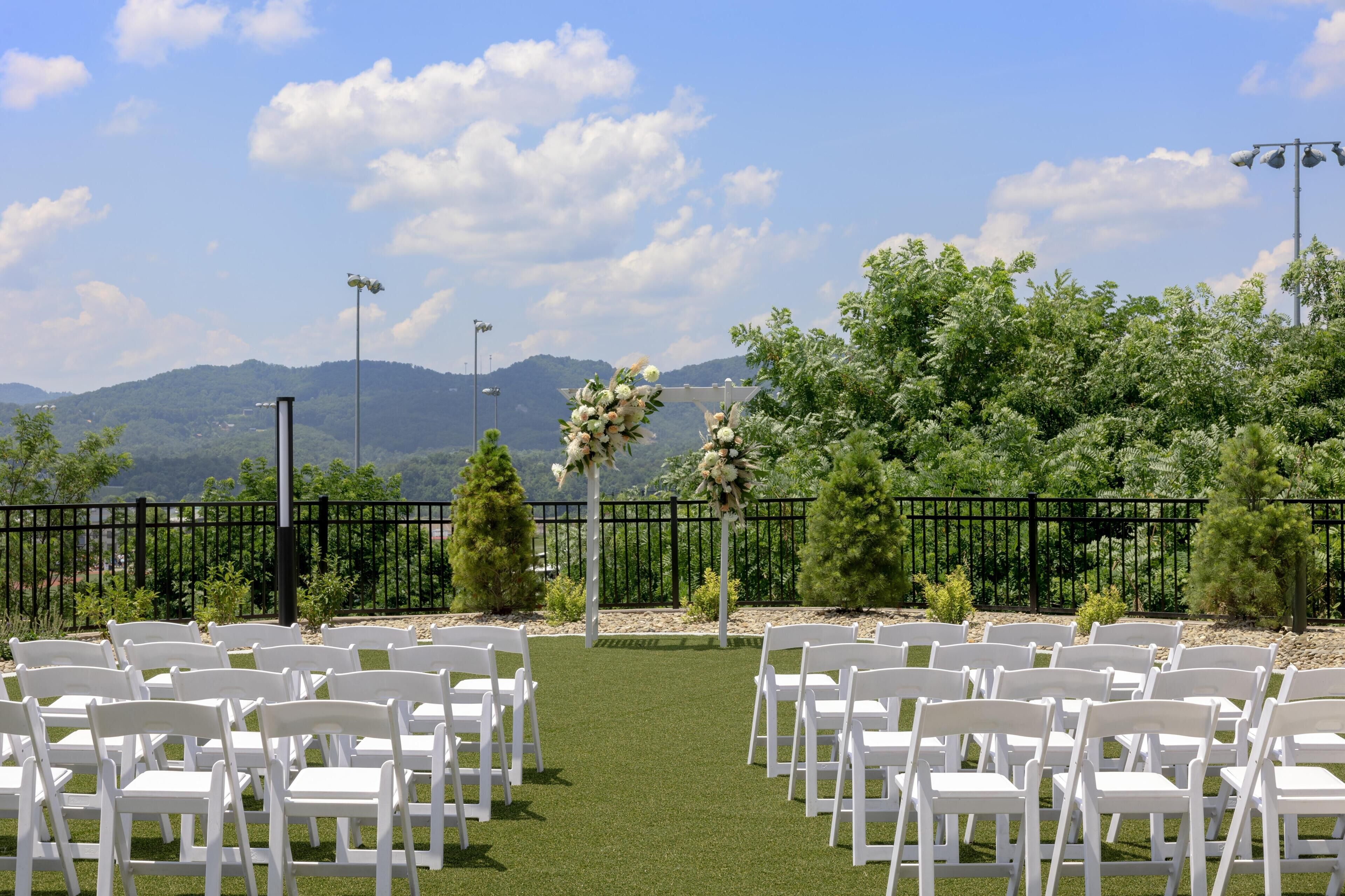 An outdoor wedding venue with white chairs arranged in rows, a black fence, and a backdrop of mountains and trees.