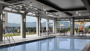A swimming pool with lounge chairs and tables, large windows with mountain views, and a covered area with umbrellas. - Tempo By Hilton Pigeon Forge (Pigeon Forge)