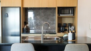 Una cocina compacta con un refrigerador de acero inoxidable, un microondas, un lavamanos con un grifo moderno y una cafetera. Hay dos sillas grises en la barra.