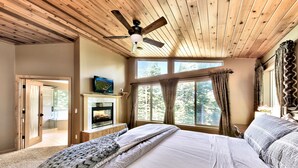 Un dormitorio con una cama grande, un ventilador de techo, una chimenea y vistas a árboles desde las ventanas.