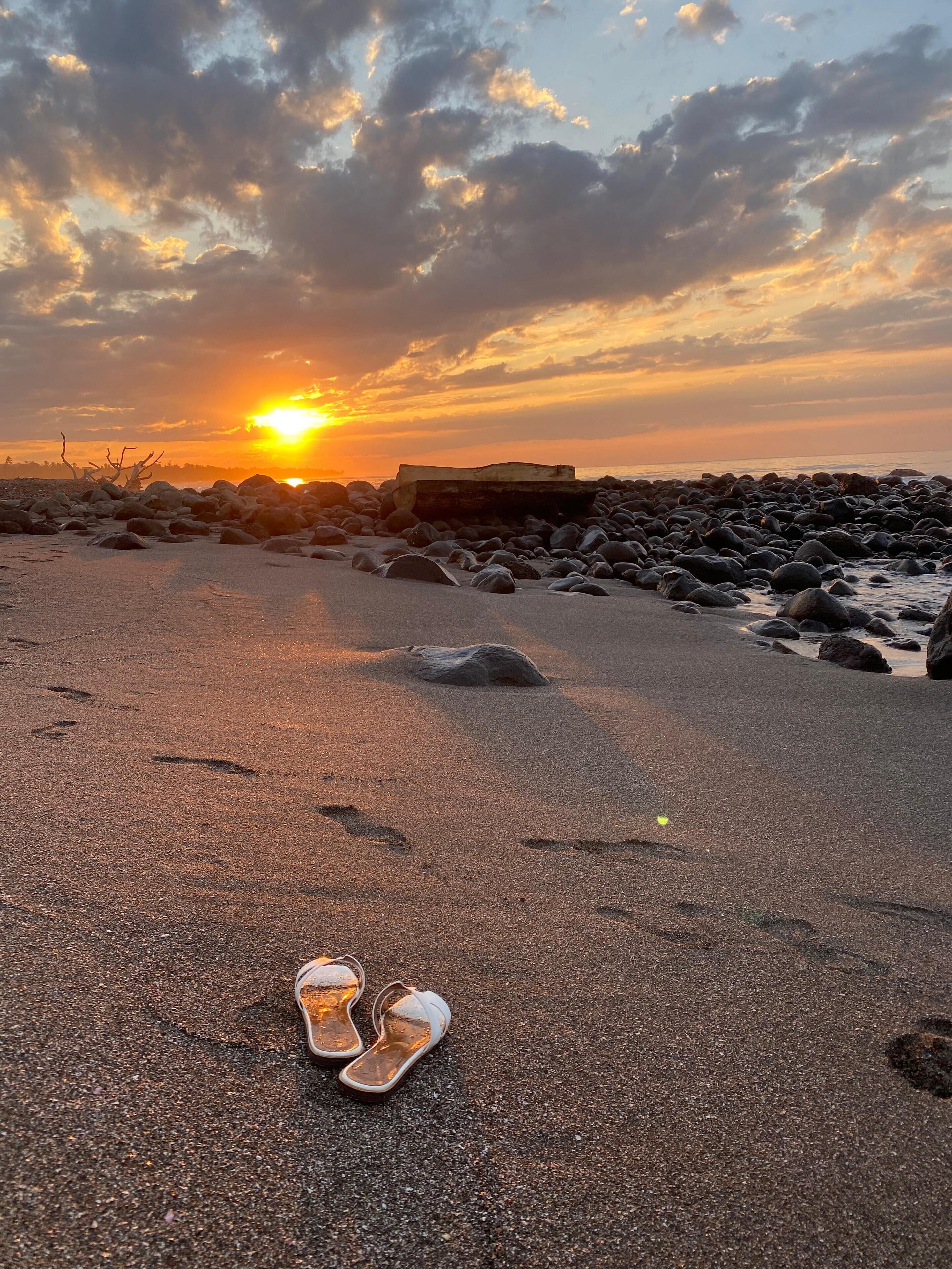 A pair of sandals on a pebbly beach with a sunset in the background.