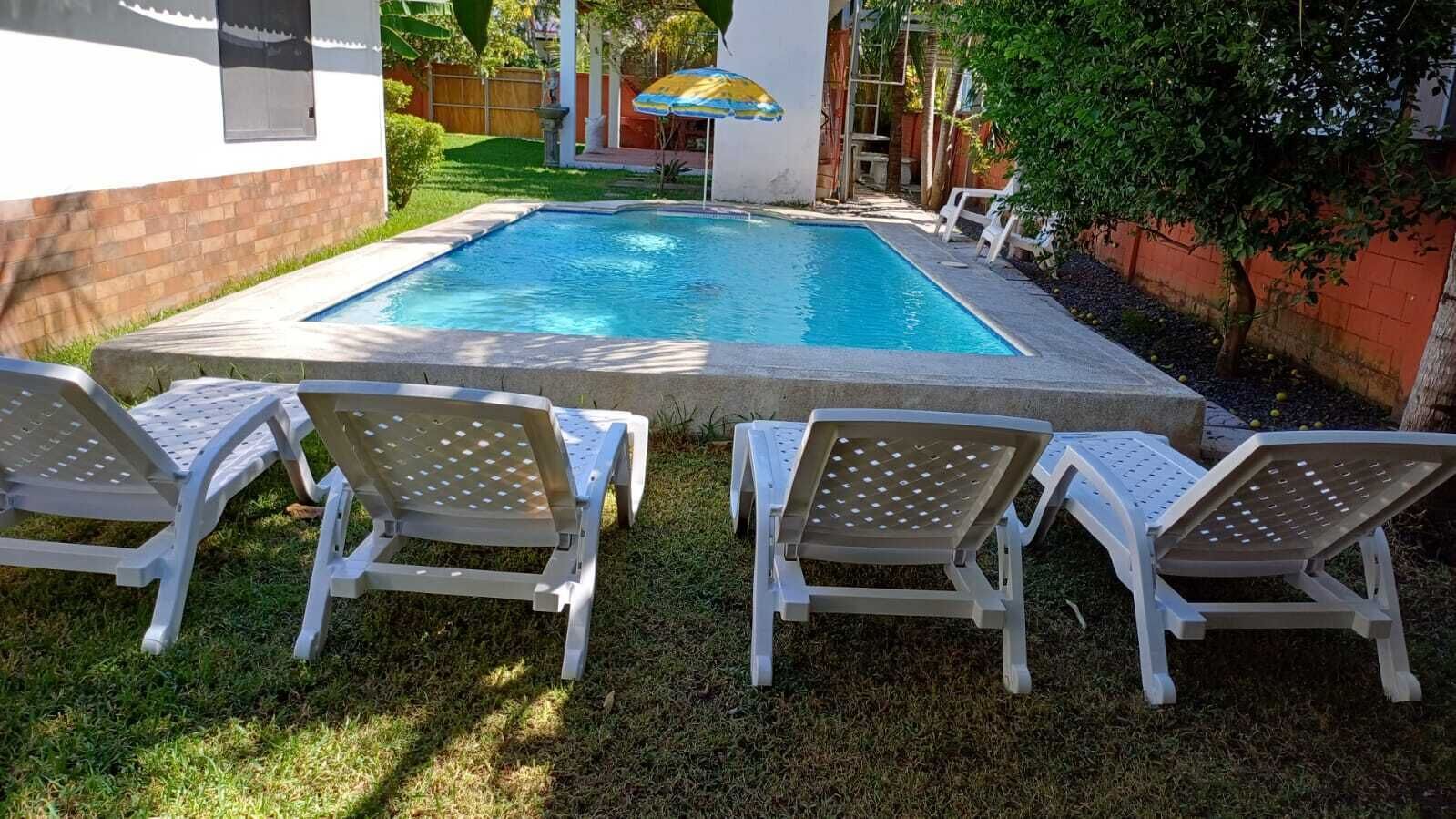 Ein Schwimmbecken mit drei weißen Liegestühlen im Vordergrund und einem Sonnenschirm im Hintergrund.