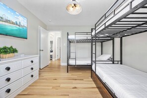 Une chambre moderne avec des lits superposés, une commode blanche dotée de poignées noires, un téléviseur à écran plat fixé au mur et un plancher en bois.