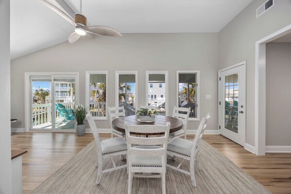 A dining area with a round table, white chairs, a ceiling fan, and large windows offering a view of palm trees and buildings.