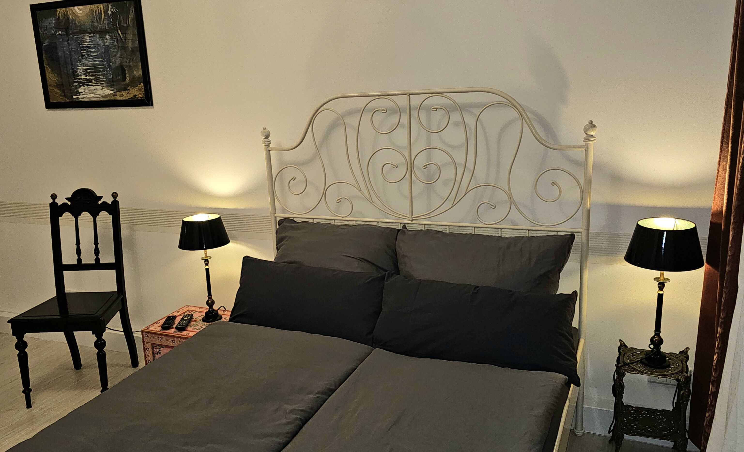 Un llit amb capçalera de metall, dues taules de nit amb làmpades, una cadira i un quadre a la paret.