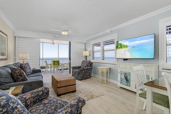 A living room with a large TV, comfortable seating, a ceiling fan, and a view of the ocean through the windows.