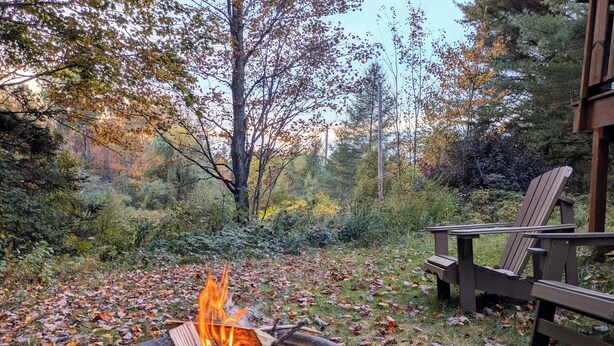 A fire pit with logs, surrounded by fallen leaves, and a wooden chair to the right. - Vintage cottage in the heart of the forest for autumn cocooning (Potton)